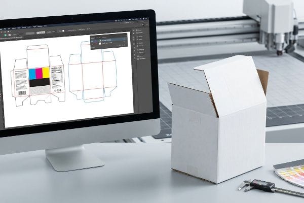 Packaging dieline with CMYK artwork and structural cut paths on a monitor, beside a white corrugated box, digital caliper, and CNC cutting machine.