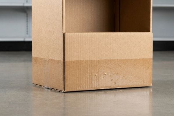 Corrugated cardboard display box bottom showing water absorption and damage from liquid exposure on a concrete floor.