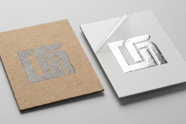 Brown kraft paper with dull silver logo next to white paper revealing shiny metallic silver foil, illustrating printing contrast.