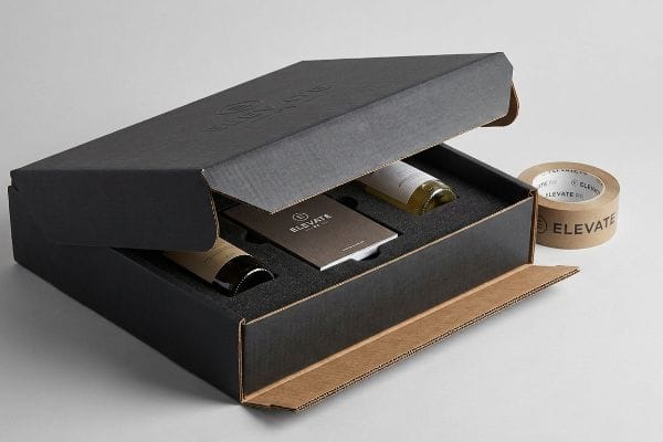 An open, structurally reinforced black PR package from Elevate, showcasing two bottles in foam inserts and a branded brown packing tape roll.