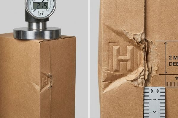 Brown corrugated box under 215 LBS pressure, showing a 2 MM deep fiber blowout near an embossed H logo, 19 MM from the crease during a Box Compression Test.