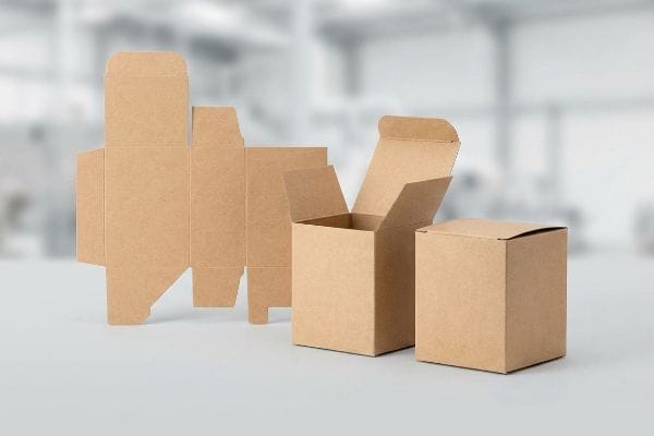 Las cajas plegables de cartón kraft muestran el proceso de ensamblaje: troquelado plano, caja abierta y caja cerrada, sobre una superficie clara.