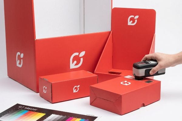 Red cardboard display boxes with a white G7 logo are measured by a colorimeter, ensuring G7 Master color calibration.
