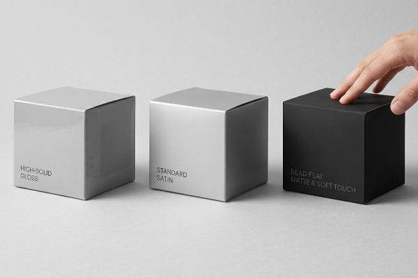Three packaging boxes demonstrating High-Solid Gloss, Standard Satin, and Dead-Flat Matte & Soft Touch aqueous coatings, with a hand touching the matte finish.