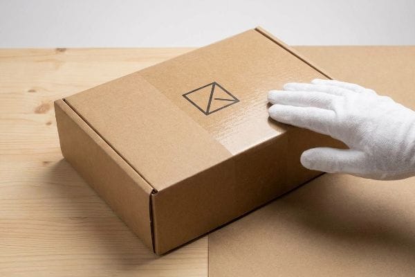 A gloved hand rests on a brown corrugated cardboard box featuring an abstract envelope logo, on a light wooden table.