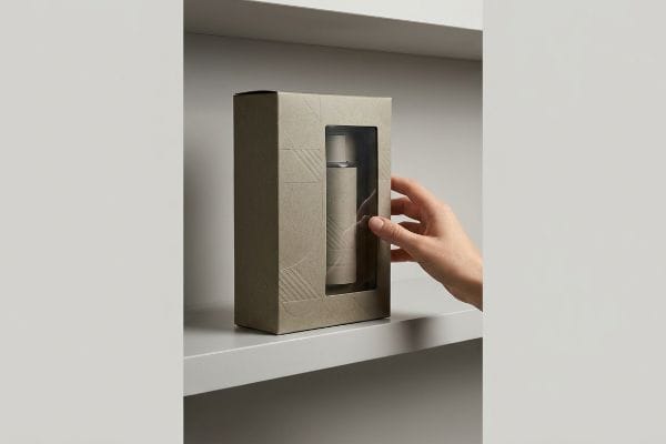 A hand reaches for a minimalist kraft display packaging box with a geometric pattern and window, showcasing a cylindrical bottle on a shelf.