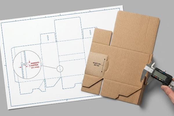 Corrugated dieline blueprint with Humidity Buffer (+1.1mm) detail, digital caliper measuring cardboard at Moisture Meter section.