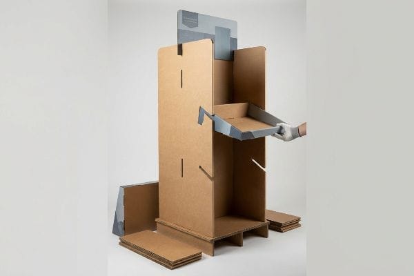 Brown corrugated cardboard retail display stand assembly, with a gloved hand inserting a gray angled shelf into an interlocking slot.