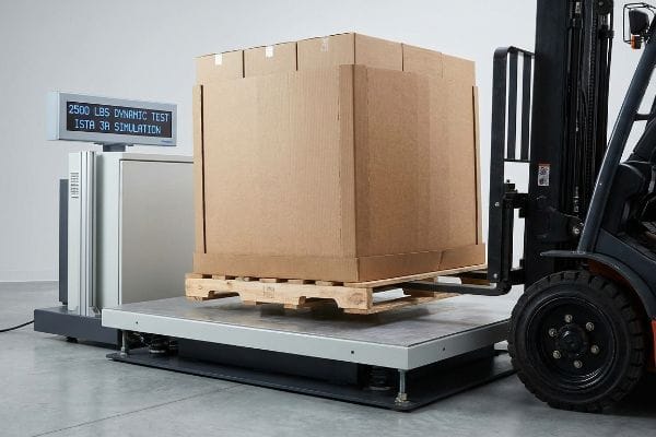 A large corrugated box on a wooden pallet is on a test platform displaying 