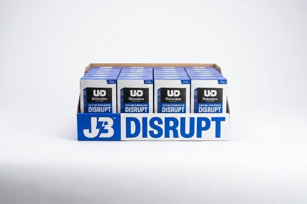 J3 Disruptのロゴが入った白い小売用ディスプレイトレイに、複数のVitaminsaneの箱が陳列されています。段ボール製のトレイには、コントラストの効いた青色のスポットカラーが施されています。.