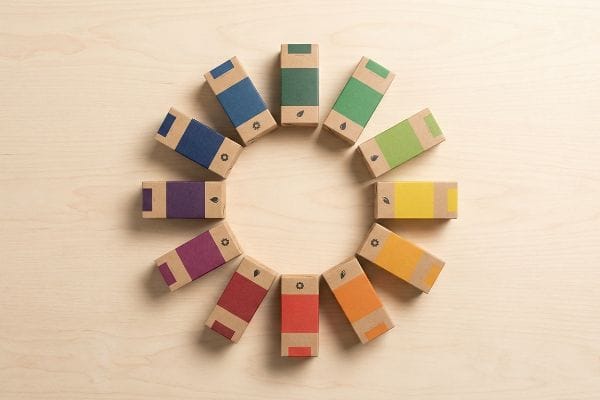 Arranged in a circular pattern, twelve eco-friendly kraft paper cardboard boxes display a vibrant retail merchandising color wheel of hues on a light wood surface.