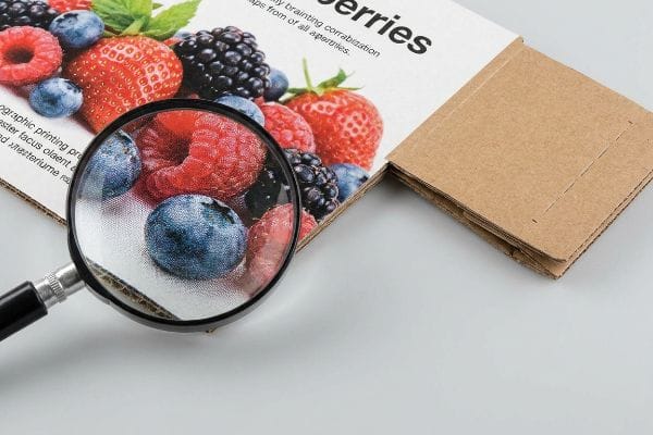 Magnifying glass shows lithographic print dots on a cardboard box with vibrant strawberries, blueberries, and raspberries.