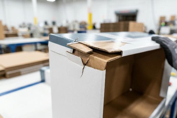 A severely torn corner of a white cardboard box with a blue top reveals cracked litho-sheet and brown corrugated board, highlighting damage from micro-friction during manual assembly.