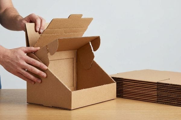 Brown corrugated display carton assembly, showing hands folding a pre-glued modular sidewall, with flat-packed PDQ trays nearby.