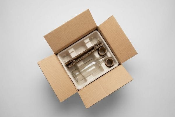 An open corrugated shipping box contains a molded pulp tray insert, securing a clear glass bottle and two cardboard rings for protective packaging.