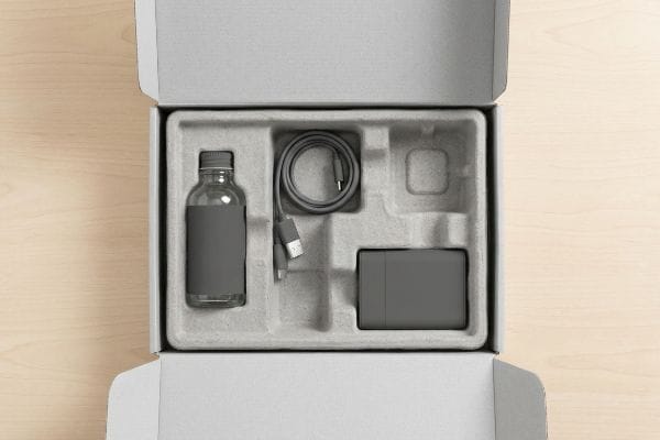 An open white cardboard box contains a gray molded pulp insert, securing a glass bottle, coiled USB cable, and power adapter for product protection.