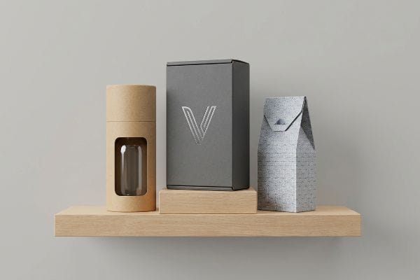 Modern product packaging display on a wooden shelf, featuring a kraft tube with a glass bottle, a dark grey box with a silver 'V' logo, and a textured light grey bag.
