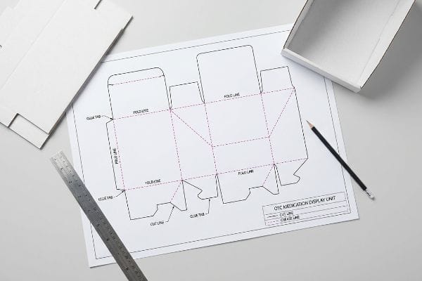Designing an Otc Medication Display Unit, a die-cut template with fold lines, glue tabs, and cut lines, alongside a ruler and pencil.