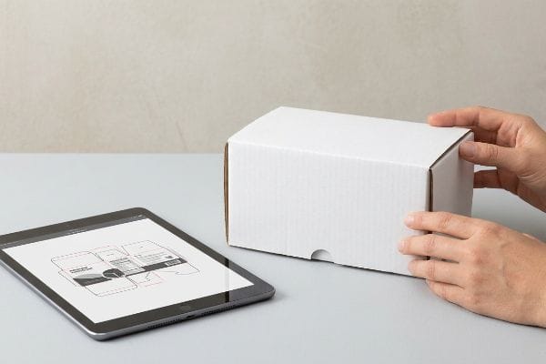 Hands inspecting a white corrugated box next to a tablet displaying a packaging dieline proof.