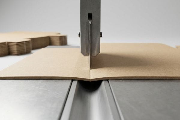 Brown paperboard being precisely creased by a steel rule die, forming a folding carton score line, preventing litho-cracking.