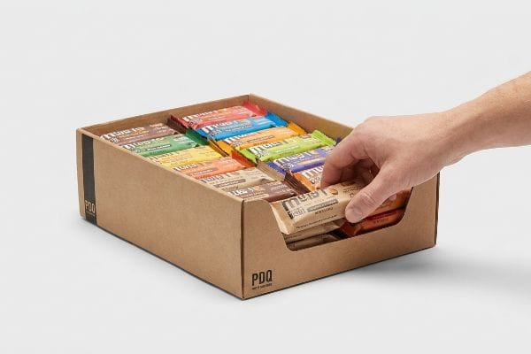 Brown cardboard PDQ display box with a hand selecting a Muesli bar, showcasing the 85% visibility rule for impulse buys.