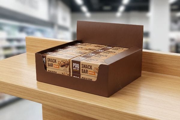 Uma caixa expositora marrom da PDQ, repleta de barras de cereais PDQ, está sobre um balcão de madeira clara, pronta para ser exposta no ponto de venda.