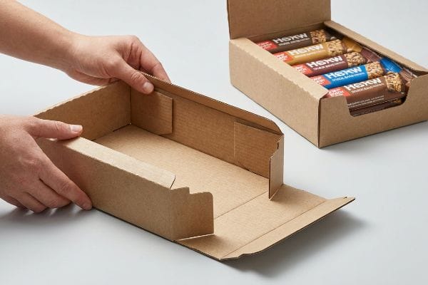 Hands assembling a kraft cardboard PDQ display tray, with a retail-ready tray of HEXW snack bars in the background.