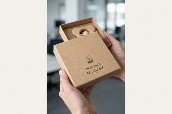 Hands hold an open brown cardboard box revealing a wooden baby teether, with
