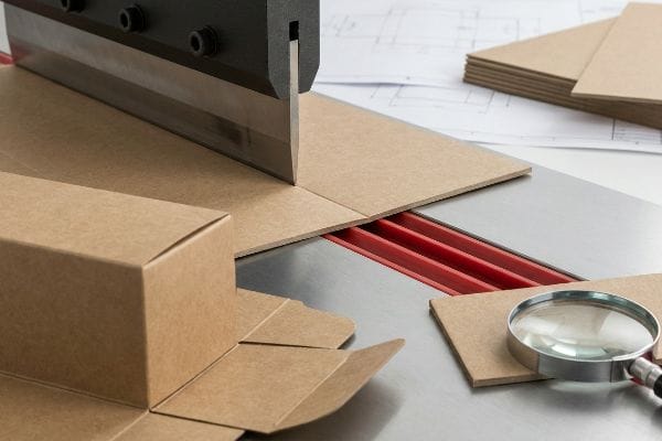 Troquel de acero para marcar cartón kraft marrón sobre una matriz de plegado de polímero rojo para una caja plegable, con una caja ya ensamblada.