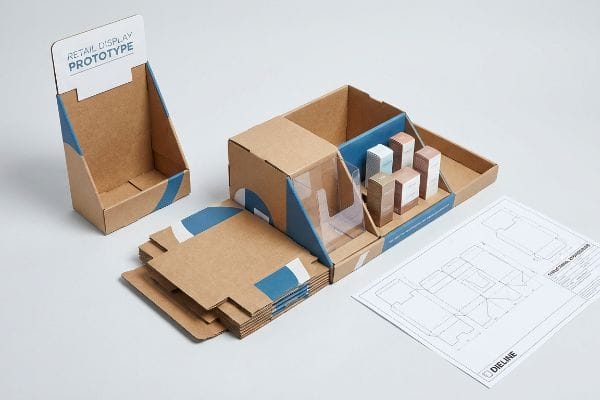 A retail display prototype, flat-packed cardboard components, and a Dieline blueprint are arranged on a white surface.