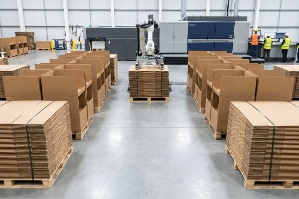An automated robotic arm stacks flat-packed corrugated cardboard displays on pallets in a high-volume production factory.
