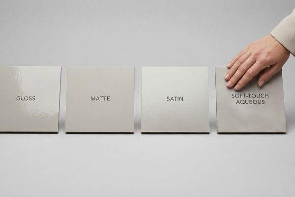 Four packaging coating samples: Gloss, Matte, Satin, Soft-Touch Aqueous, with a hand touching the soft-touch card.