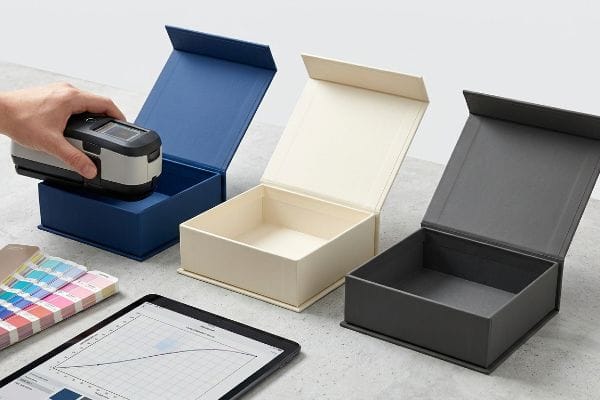 Spectrophotometer measures dark blue rigid box color accuracy. Cream, grey boxes, color swatch, and tablet with prepress data visible.