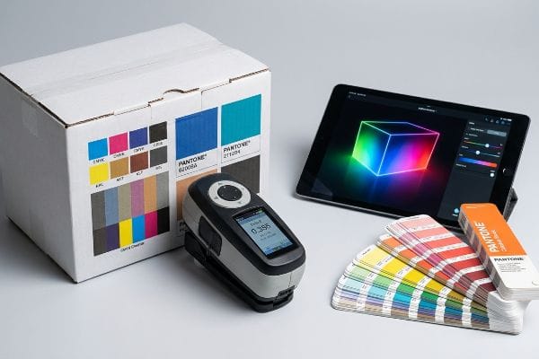 White box with CMYK, Pantone swatches, spectrophotometer showing Delta-E, iPad with RGB cube, and Pantone Color Bridge fan decks.