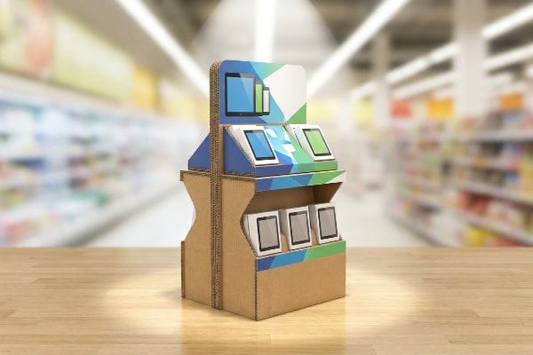 A multi-tiered corrugated retail display, featuring vibrant spot-color graphics and six white tablets with blue and green screens, stands on a wooden surface in a blurred supermarket.