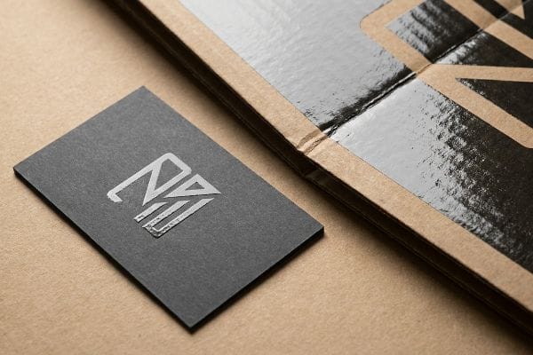 Matte dark grey business card with a silver spot UV logo, placed beside a corrugated cardboard display featuring glossy black print on brown kraft.
