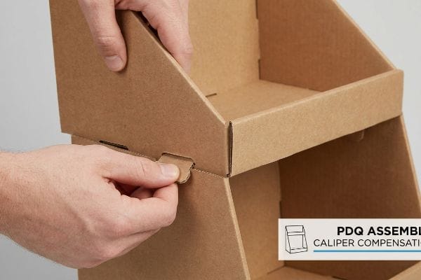 Brown corrugated cardboard PDQ display assembly, a hand inserting a tab into a slot, highlighting Caliper Compensation for frictionless fit.