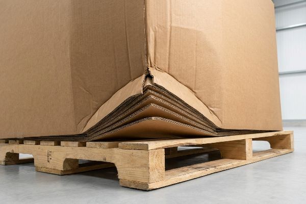 Triple-wall corrugated cardboard box corner overhanging a wooden pallet, revealing multiple fluted layers and linerboards.