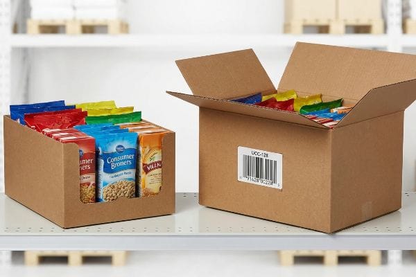 Brown cardboard display tray and shipping box filled with colorful snack bags, featuring a UCC-128 barcode and Consumer Broners chickpeas.