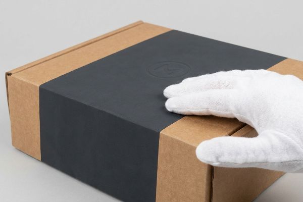 Soft touch aqueous coating on a corrugated box, gently touched by a white-gloved hand, highlighting the velvet-like surface and subtle embossed logo.