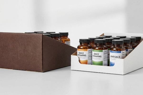 Two corrugated cardboard display trays, one brown and one white, filled with amber Daily Vitamins and Omega-3 bottles, featuring low-profile front lips for pharmacy products.