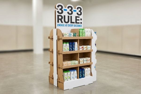 段ボール製のエンドキャップディスプレイには、「3-3-3ルール：あらゆる距離で積極的に関わろう」というメッセージが掲げられており、シャンプーや石鹸製品が陳列されている。.