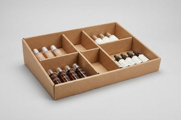 Brown corrugated cardboard display tray with modular dividers, displaying asymmetrical clusters of small cosmetic bottles: light, white, dark, and dropper types.