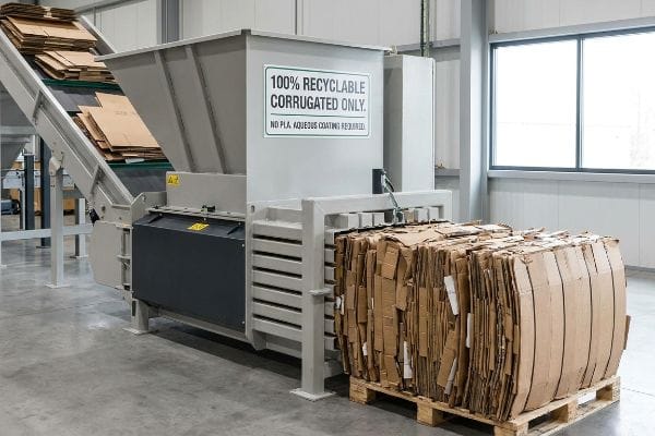 Corrugated cardboard boxes feed into a grey industrial baler, displaying a sign: '100% Recyclable Corrugated Only. No PLA. Aqueous Coating Required.' A compressed bale rests on a pallet.