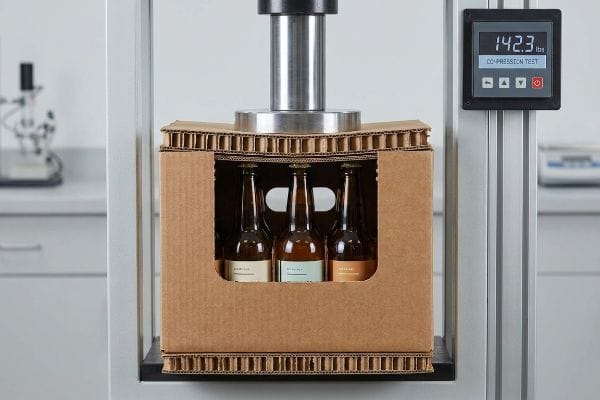 Corrugated cardboard 6-pack with glass bottles under compression test, digital gauge reads 142.3 lbs.