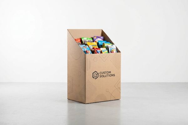 A freestanding corrugated retail dump bin, branded with Custom Solutions logo and geometric patterns, holds assorted snack bags including M&M's.