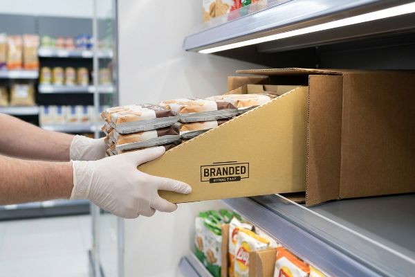 A Branded Hymal Tray of pre-packed bread rolls is placed on a retail shelf by gloved hands, demonstrating efficient PDQ tray restocking.
