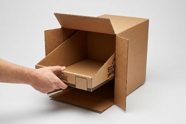 A hand pulls a Brandic PDQS display tray from a corrugated master carton, demonstrating the quick-release offset.