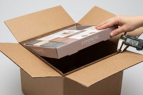 Digital caliper measuring 0.250 inches clearance between a brown corrugated shipping box and a 'Cosmetic' product tray, illustrating geometric offset tolerance for smooth extraction.