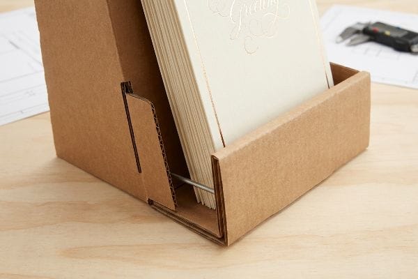 Corrugated cardboard greeting card display with a 3mm steel support bar reinforcing the front lip, holding foil-stamped 'Greeting' cards.